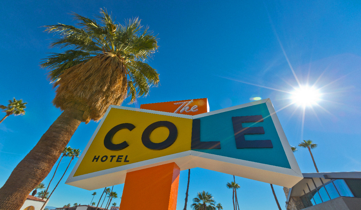 A colorful sign reading 'The Cole Hotel' in front of a palm tree and a building under a bright sun.